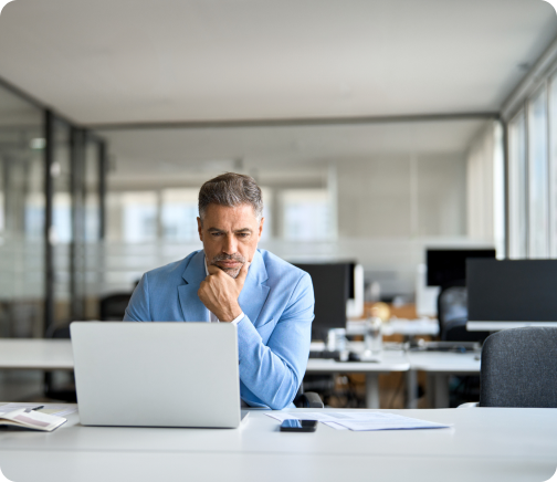 A thoughtful man in a blue blazer sits at a desk in a modern office, looking at his laptop.